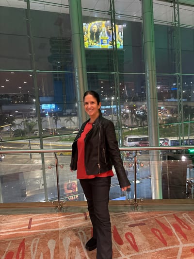 A woman is standing in front of a large glass window in what appears to be an interior space, possibly a lobby or mezzanine. The window provides a view of a city at night. The woman is wearing a black leather jacket over a red top, black pants, and black shoes. The floor has a patterned carpet. Reflected in the glass window are buildings, streets with traffic, and other lights, suggesting the location is urban. A sign, possibly a movie poster, is partially visible on one side of the window. The reflections and the illuminated urban landscape through the glass suggest the scene is set in the city of San Borja, Peru.
