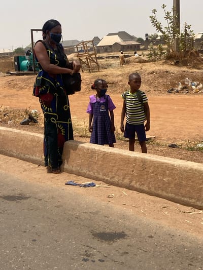 The image displays an outdoor scene in an unspecified urban or semi-urban area of Nigeria. The setting features a segment of asphalt road in the foreground, bordered by a concrete curb separating it from an elevated dirt shoulder. Scattered debris, including a blue fabric item and light-colored fragments, rests on the dirt adjacent to the curb.
Three individuals are present on the dirt shoulder. On the left, an adult female with dark skin stands wearing a long, patterned dress featuring blue, black, yellow, and red/orange motifs. She wears dark brown sandals, large gold-colored hoop earrings, and a dark face mask covering her mouth and nose. She holds a dark bag in her left hand. To her right stands a young girl, approximately 5-7 years old, also with dark skin, wearing a purple and dark blue plaid school pinafore over a white-collared shirt, forming a school uniform. Her hair is styled in two dark buns. A dark face mask covers her mouth and nose. To the right of the girl is a young boy, approximately 6-8 years old, with dark skin and short dark hair. He wears a green and white horizontal striped t-shirt, dark blue shorts, and light-colored sandals, with a light-colored backpack on his shoulders. All three individuals are standing upright, facing generally forward or slightly to the right of the viewer's perspective.
The background consists of dry, reddish-brown earth and sparse, dry vegetation. To the far left, a large green industrial generator unit is visible. Further back, various modest and partially constructed buildings, likely residential or commercial, are scattered across the landscape. These structures appear to be made of concrete blocks or similar materials, some with light-colored walls and sloped roofs. A wooden scaffolding-like structure is also visible. In the distant background, several indistinct figures can be observed near buildings and a large mound of earth or rock. The sky is hazy and light-colored, suggesting dry and possibly dusty atmospheric conditions.