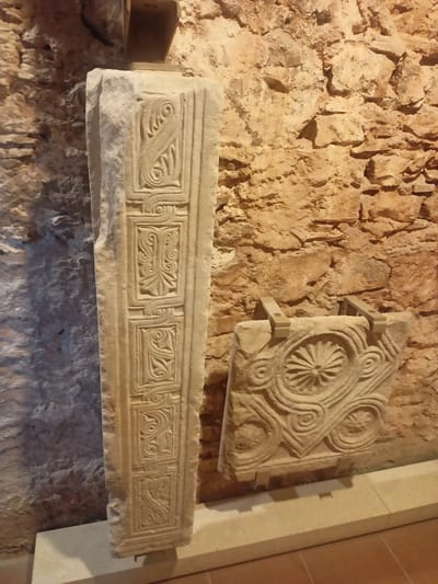 In the Municipal Unit of Anthidon, Greece, an archeological display features two stone artifacts against a rough, tan stone wall. The artifact on the left is a tall, rectangular stone pillar, standing upright. Its surface is divided into rectangular panels, each containing intricate carvings of scrolling patterns and stylized plant-like motifs. The artifact on the right is a smaller, squarish stone block, positioned horizontally. It is adorned with a central circular design resembling a flower or starburst, surrounded by swirling, curvilinear patterns. This artifact is held in place by two light-colored, angled brackets. The floor in front of the artifacts appears to be tiled in a reddish-brown color. The lighting suggests an indoor environment, possibly a museum or exhibition space.