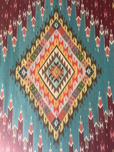The image shows a colorful rug featuring a large diamond-shaped design in the center. The diamond is composed of concentric shapes using geometric patterns and a variety of colors including red, yellow, brown, grey, and blue. The background is teal with additional rows of geometric motifs near the top and bottom, colored with maroon. The designs have a Southwestern or Native American textile appearance.