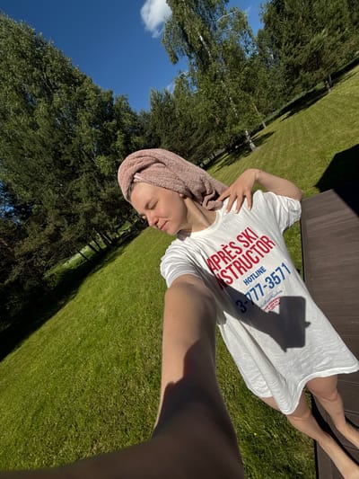 An outdoor selfie depicts a person, appearing female, situated on a dark brown slatted wooden surface in Копнино, Russia. The individual has a light pink towel wrapped around their head and wears an oversized white t-shirt. The t-shirt prominently displays "APRÈS SKI INSTRUCTOR" in red text, "HOTLINE 3-777-3571" in blue, and smaller text reading "SkiBum Wisdom YOUR PERSONAL GUIDE TO APRÈS FUN". The person's left hand, adorned with light-colored nail polish, rests on their neck. Their head is tilted, and eyes are partially closed. Bare legs and feet are visible on the right. The background features an expansive, brightly sunlit green lawn, bordered by a dense line of deciduous and coniferous trees casting long shadows. Above the tree line, a clear blue sky with sparse white clouds is visible.
