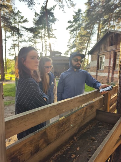 Three individuals are gathered outdoors near a wooden structure in Онохино, Russia. In the foreground, a woman with long brown hair, wearing a dark striped shirt, looks to her left. Behind her and to her right, another woman with long brown hair and sunglasses, dressed in a dark vest over a light-colored shirt, looks downwards. A man wearing a dark hooded jacket, sunglasses, and a baseball cap is on the far right, holding a smartphone in his right hand. He is looking towards a wooden trough filled with dry leaves and pine cones. The background features a rustic wooden building with a brick lower section, surrounded by tall pine trees. The scene is illuminated by the warm light of the sun, suggesting it is late afternoon or early evening. The environment appears to be a rural or wooded area.