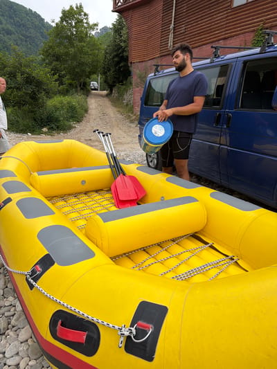 A man with a beard and short dark hair, wearing a navy blue t-shirt and black shorts, stands next to a bright yellow inflatable raft. He is holding a blue plastic bucket with white lettering. The raft is partially inflated and rests on a rocky surface. Two red and silver paddles are positioned on top of the raft. In the background, a blue van is parked next to a weathered wooden building with a red roof. A dirt road leads away from the scene, bordered by lush green trees and vegetation. In the distance, a white SUV is visible on the road, and rolling hills covered in trees rise against a cloudy sky. The overall scene suggests an outdoor activity, possibly related to water sports, taking place in a rural or semi-rural environment. There is no specific indication of the time of day or weather, but the overcast sky suggests it could be an unpredictable day. The location cues, such as the wooden building and the natural landscape, point towards a natural setting like a riverbank or lake shore. The man's posture and the presence of the raft and paddles indicate preparation for or participation in an activity.