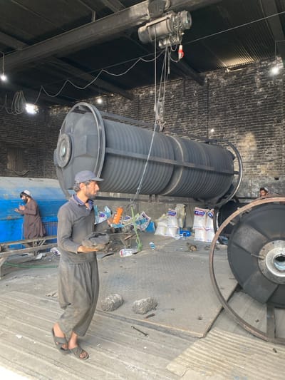 This image captures an interior view of an industrial workshop in Daman, Afghanistan. The scene features three males amidst various heavy machinery and components under a corrugated metal roof with exposed brick walls.
In the foreground, a man with a beard and a dark cap wears a dark long-sleeved shirt, dark pants, and sandals. He is walking towards the viewer's left, carrying two dark, irregularly shaped objects, possibly machine parts or materials.
Dominating the central mid-ground is a large, dark grey cylindrical industrial object, approximately 4-5 meters in length, with a corrugated exterior, supported by a metal frame. This object is positioned directly beneath an overhead electric hoist system, which is mounted on a steel beam across the ceiling. Cables and a hook from the hoist extend towards the cylindrical object, suggesting it is either being moved or prepared for movement.
To the left of the central cylindrical object, a second man, dressed in a brown thobe and a white prayer cap, is visible, partially obscured. He stands next to a blue industrial machine or tank, facing away from the camera, with his hands clasped, appearing to be engaged in prayer or deep contemplation.
To the right, another large, dark metal wheel or disk-shaped component lies on the floor, suggesting it is either being worked on or awaiting assembly. A third male is partially visible in the background behind this component.
The floor is a mix of concrete and metal plating. Scattered across the floor are various items, including white sacks labeled "SECO," tools such as wrenches and a screwdriver, and miscellaneous debris. Exposed incandescent light bulbs hang from the ceiling, providing illumination. The overall environment suggests an active industrial fabrication or assembly setting.