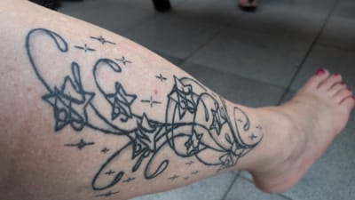 The image shows a close-up of a person's left leg with a tattoo of swirling vines and stars. The tattoo is black ink and covers a significant portion of the calf. The skin appears fair with some freckles and minor blemishes. A bare foot with pink nail polish is visible on the right side of the frame, suggesting the person is likely outdoors or in an open area. The background consists of gray tiled flooring. The overall impression is of an ordinary day in a public or outdoor space. The image does not contain any specific visual cues that would allow for definitive identification of the location as Dissen am Teutoburger Wald, Germany.