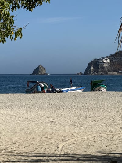 A man is standing on a sandy beach next to a jet ski. The jet ski is white and blue with some red accents. The man is wearing a dark t-shirt and shorts. The beach is made of light-colored sand. In the background, there is blue water and a rocky cliff with some buildings on top. The sky is clear and blue. There is a small boat with a green cover in the water near the shore. The scene appears to be a sunny day, likely in the morning or afternoon given the shadows. The location is identified as Santa Marta, Colombia, suggesting a tropical coastal environment. The overall mood is relaxed and recreational. The jet ski has visible text that reads "CP-16-ZM".