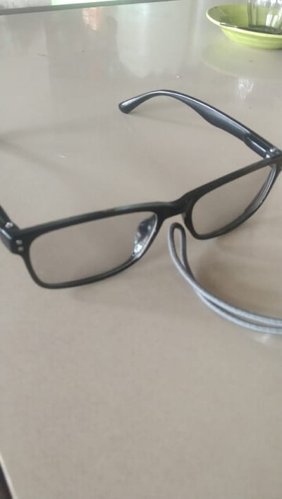A pair of black-rimmed eyeglasses with clear lenses are placed on a light-colored, smooth surface, possibly a table, in what appears to be an indoor setting. The eyeglasses are positioned at an angle, with the left lens facing forward and the right lens slightly obscured. A thin, gray cord, possibly a glasses strap, is visible looping around the right temple of the glasses. In the upper right corner of the image, a portion of a green dish with a clear glass or cup is visible. The lighting suggests a well-lit environment, with reflections visible on the lenses and the surface. The overall scene is static, with no individuals or overt actions depicted. The location is Meunasah Pupu, Indonesia.