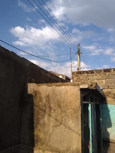 The image displays an outdoor urban environment within Njoro ward, Kenya, under a blue sky with scattered white cumulus clouds. Direct sunlight illuminates the scene, casting distinct shadows. In the foreground, a weathered, light brown concrete wall with visible discoloration and a prominent crack in its lower right quadrant occupies the left side. Behind this, a darker concrete wall, topped with sparse vegetation, is partially visible. To the right, a wall constructed from rough-hewn stone blocks rises. A wooden utility pole, heavily laden with numerous black wires, stands above the stone wall in the upper right quadrant. Multiple black utility wires traverse the sky horizontally and diagonally, connecting to this pole. A slender metallic rod or antenna is also present near the utility pole. In the lower right, a doorway with a light blue metal frame and a partially open, matching light blue door or shutter is visible, topped by a small concrete overhang. A crumpled white object rests on the stone wall directly above the doorway. No individuals or specific actions are depicted.