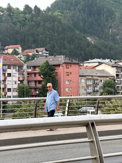 The image depicts a street scene in Konjic, Bosnia and Herzegovina, characterized by a man walking on a pedestrian bridge. The man, an elderly Caucasian with white hair and sunglasses, is wearing a light blue short-sleeved collared shirt and dark pants. He is walking towards the right of the frame, with his left foot forward. The bridge has a metal railing in the foreground.
Behind the bridge, a dense urban landscape unfolds, dominated by multi-story residential buildings of varying architectural styles and colors, including pink, beige, and pale pink. The buildings feature multiple windows, balconies, and some visible air conditioning units. Trees and greenery are interspersed among the buildings, and a lush, densely forested hillside rises in the background, suggesting a location at the base of a mountain or significant incline. A white van and a dark-colored vehicle are visible on the road beneath the bridge. A flag is visible on a pole to the far left of the frame, partially obscured by a building. The sky is overcast.