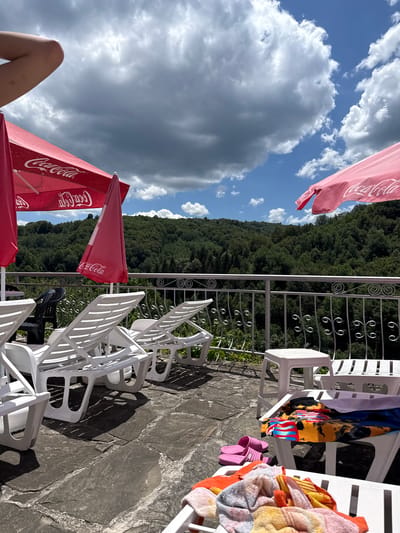 This image depicts an outdoor scene at a resort or hotel in Shipkovo, Bulgaria, likely a spa or pool area. In the foreground, several white plastic lounge chairs are arranged on a flagstone patio. Some of the chairs have towels draped over them, and a pair of pink flip-flops are visible on one chair. In the middle ground, a white metal railing with decorative elements separates the patio from a steep, densely forested hillside that dominates the background. Two large red Coca-Cola branded umbrellas are partially visible in the upper left and upper right portions of the frame, providing shade. The sky above is a mix of blue sky and dramatic, heavy grey clouds, suggesting variable weather conditions. A person's arm is visible in the upper left corner of the image, suggesting a human presence, though no other individuals are clearly depicted. The overall impression is one of a scenic, possibly relaxing, outdoor setting in a natural environment.