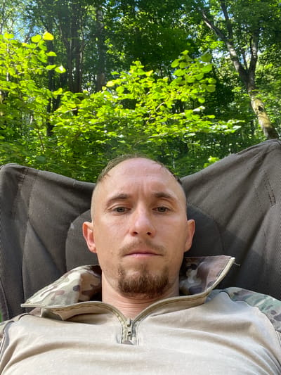 A man, with light skin, a shaved head, and a goatee, lies on his back on a dark, textured surface. He is wearing a camouflage-patterned jacket with a zipper that is partially open, revealing a light-colored shirt underneath. His head is tilted slightly upwards and to his left. The man's expression is neutral, with his eyes open and looking towards the camera.

Behind the man, a dense canopy of green leaves and tree branches is visible, suggesting an outdoor setting in a forest or wooded area. The sunlight filters through the leaves, creating dappled patterns and highlighting the vibrant green of the foliage. The overall impression is one of a relaxed moment spent outdoors during the daytime. The image appears to be taken in Nikolayevka, Russia. No text is visible in the image.