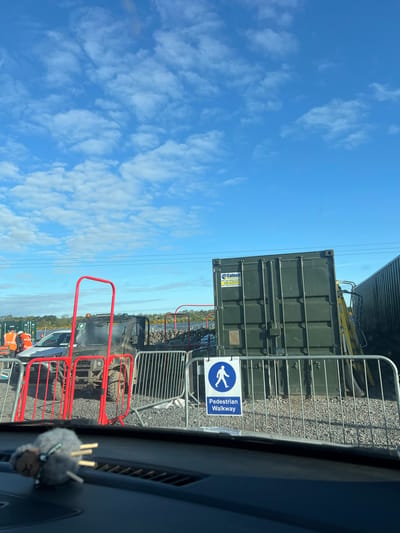 A view, potentially from inside a vehicle, shows a construction or industrial site. A green shipping container is positioned near a pedestrian walkway sign, with metal fencing nearby. A red barrier is in the foreground, and construction or utility vehicles and workers are visible further into the site. The sky is blue with scattered clouds. The location is Grange Ville, Ireland.