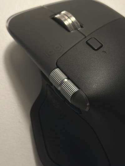 A black Logitech MX Master 3S wireless mouse is placed on a white surface. The mouse is angled, showing its top and right side. Key features visible include the scroll wheel with a metallic finish and intricate ridges, a small rectangular button, and a textured grip on the right side. The Logitech logo is subtly embossed on the mouse's surface. The lighting casts a soft shadow to the left of the mouse. The scene depicts a product detail shot, typical of e-commerce photography. There is no indication of the city of Kfar Saba, Israel, or any other location within the image.