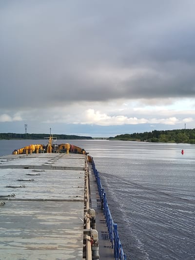 The image shows a view from a ship moving on a body of water, likely a river or wide canal, with land visible on both sides. The ship has a large, flat deck with gray, weathered surfaces and yellow equipment towards the bow. A blue railing runs along the starboard side of the ship.
The water is dark with ripples and wakes indicating movement. In the distance, a red buoy marks the channel. The land is covered in green trees. A power line tower is visible on the left side of the image.
The sky is overcast with gray clouds. The image likely depicts a view from near the city of Нифантово, Russia.