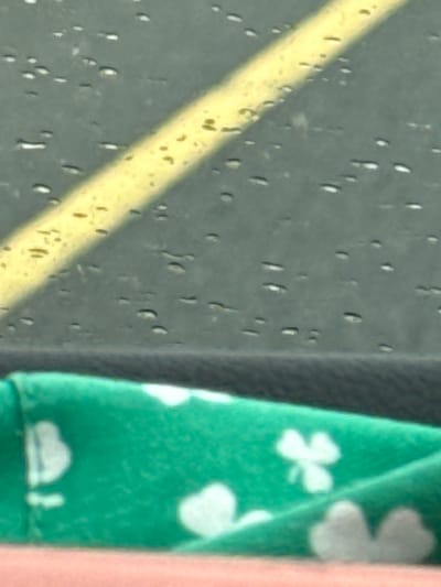 The image shows a close-up of a surface that appears to be a window with water droplets. A yellow line is visible on the surface, possibly a lane marker on a road viewed through the window. Below the window, a green fabric with white clover-like patterns is visible, suggesting it is a decorative item or clothing. The bottom of the image shows a pink surface, likely a hand or another object. There is no discernible information about Father Campion Place, Ireland within the image.