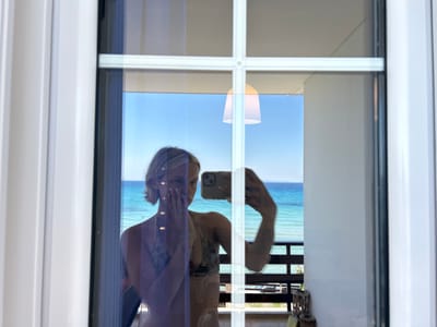 A woman, reflected in a window pane, is taking a selfie with her phone. She is wearing a bikini and has her hand over her face. The reflection shows a balcony and the sea beyond, suggesting a coastal location. The window frame is white and there is a faint reflection of a lamp. The scene appears to be in Arkadii Municipal Unit, Greece, a region known for its beautiful coastline.