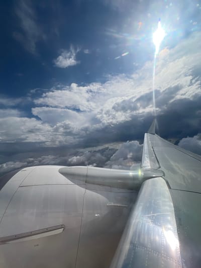 The image depicts an airplane wing and engine in flight, with a backdrop of clouds and a bright sun. The wing is silver and reflective, with visible rivets and other details. The engine is white and dominates the right side of the frame. The clouds vary in color and density, ranging from dark and stormy to light and fluffy. The sun is positioned high in the sky and casts a strong glare. The location in Rustavi, Georgia, is not discernible from the image.