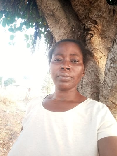 A woman stands close to the camera, her face centered and filling the frame. She has dark skin and wears a light-colored, plain T-shirt. Behind her, a large tree trunk dominates the background, its bark textured and detailed. To the left, foliage and branches extend into the frame, with a hint of a rural landscape visible in the distance. The lighting is bright, but somewhat harsh, casting shadows on the woman's face and highlighting the tree's details.