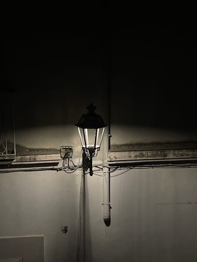 This is a photograph of an outdoor lamppost at night, likely in Santa Maria a Vico, Italy. The lamppost, with a black metal frame and clear glass panes, is illuminated, casting a bright light on the textured, off-white wall behind it. The wall appears aged and shows signs of discoloration and some structural elements like pipes and wiring are visible. The upper portion of the image is dark, suggesting the night sky or a dark facade above. There are no people or animals visible in the frame. The scene is devoid of any overt activity, presenting a static, atmospheric depiction of a dimly lit urban environment.