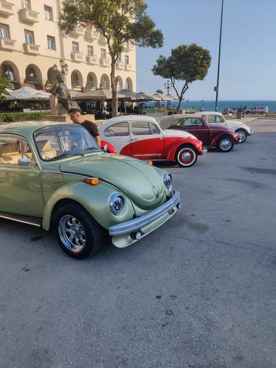 Four classic Volkswagen Beetles are parked in a line on an asphalt surface. The front Beetle is a light green, followed by a red and white convertible, a burgundy convertible, and a white Beetle. A man in a black t-shirt is standing near the open driver's side window of the green Beetle, looking down. In the background, a large, light-colored building with arched doorways and balconies lines a street. Trees, streetlights, and outdoor seating areas with umbrellas suggest a public promenade or waterfront area. Beyond the cars and building, the blue sea is visible, with several ships on the horizon. The sky is clear and blue, indicating a daytime setting with pleasant weather. The overall scene evokes a sense of nostalgia and appreciation for vintage automobiles in a picturesque, possibly tourist-oriented, location. The text "www.cosmorent.gr" is visible on the side of the red and white Beetle.