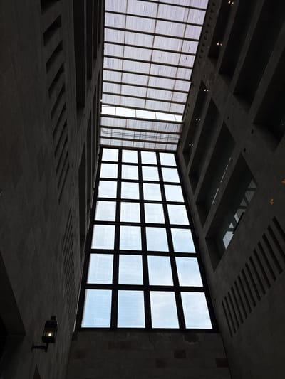 The image shows an interior space with a high ceiling and two large window structures. One window structure is composed of thin vertical slats, while the other is made of square panes. The walls and ceiling are constructed from a textured, light-colored stone. The ceiling has rectangular indentations and a rectangular skylight. On the floor, there are rectangular recesses, and in the lower right corner, a small light fixture is visible. The image provides no information about the location in Ciudad López Mateos, Mexico.