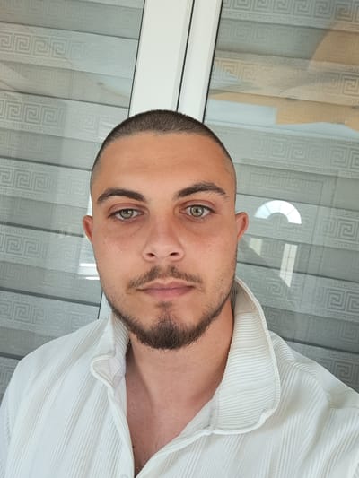 A close-up, head and shoulders selfie of a man with a buzz cut and a beard, wearing a white, textured collared shirt. The man is looking directly at the camera with his head slightly tilted. His eyes are green. The background consists of a window with horizontal blinds featuring a Greek key pattern. A reflection of a curved window frame is visible on the blinds. The setting appears to be indoors, with the lighting suggesting daytime. This scene is not located in the city of Ignatievo, Bulgaria.