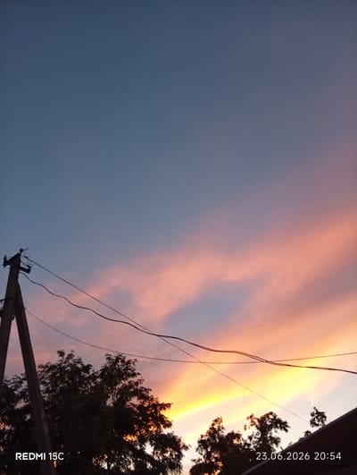 This is a vertical, outdoor, mid-range shot of a sky at dusk. The sky is a gradient of colors, transitioning from a deep blue at the top to soft pinks and oranges nearer the horizon. The colors suggest it is either sunrise or sunset. Silhouetted against the sky are the dark shapes of trees and a wooden utility pole with wires. The utility pole is on the left side of the frame and appears to be made of rough, weathered wood. Several electrical wires stretch across the frame from the pole, disappearing out of view. The trees are dense and leafy, also silhouetted. The overall mood is peaceful and serene, evoking the quiet transition between day and night. Visible text in the bottom left corner reads "REDMI 15C," and in the bottom right corner, the date and time "23.06.2026 20:54" are displayed. The location is Mykolaivka, Ukraine. The weather appears clear, with no visible clouds other than the wisps catching the colorful light.