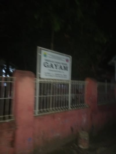 The image depicts a low-light, blurry outdoor scene, likely captured at night. A red-painted concrete wall spans horizontally across the lower and mid-ground, topped by a white metal fence composed of vertical bars. A prominent rectangular white sign is mounted on the white fence. The sign displays multi-colored logos and legible text, including "PEMERINTAH KABUPATEN JEMBER" (Government of Jember Regency), "DINAS KEBUDAYAAN DAN PARIWISATA" (Department of Culture and Tourism), and the large word "GAYAM". Further text below "GAYAM" details "JL. RAYA GAYAM DSN. CANGAR DESA GAYAM KEC. RAMBIPUJI KAB. JEMBER", identifying the specific location as Gayam Village, Rambipuji District, Jember Regency, Indonesia. Dark, indistinct foliage is visible behind the fence and sign, suggesting an overgrown area. A small, partially obscured cylindrical object, possibly a container with red and yellow markings, rests on the dark ground at the base of the red wall on the right side. No individuals are present.