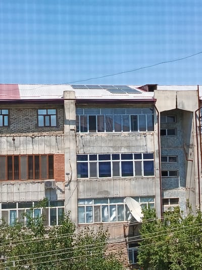 The image shows the exterior of a multi-story apartment building located in Nukus, Uzbekistan. The building appears to be constructed of concrete and brick. The facade features numerous windows, some with glass panes and others with darker coverings. There are also balconies and solar panels installed on the roof. A satellite dish is visible mounted on the side of the building. Trees and power lines are present in the foreground, suggesting an urban environment. The sky above is a clear blue.