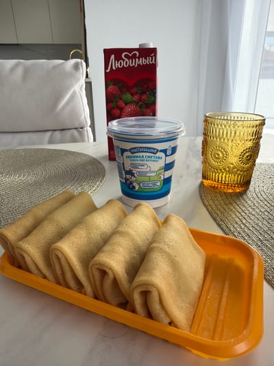 Here is a forensic description of the image:
The image shows a table setting with various food items, likely in a home in Молодёжный, Russia.
**Objects:**
* A stack of folded pancakes in an orange serving dish.
* A container of sour cream labeled "Prostokvashino" (in Russian).
* A carton of strawberry juice labeled "Lyubimy" (in Russian).
* A decorative amber-colored glass.
* Two woven placemats.
* A portion of a white cushioned chair is visible.
**Environment:**
* The setting appears to be indoors, likely on a table near a window, indicated by the presence of sunlight and window frame in the background.
**Summary:**
The image depicts a breakfast or brunch scene with pancakes, sour cream, and strawberry juice. The Russian labels indicate the location is in Russia, possibly in a home.