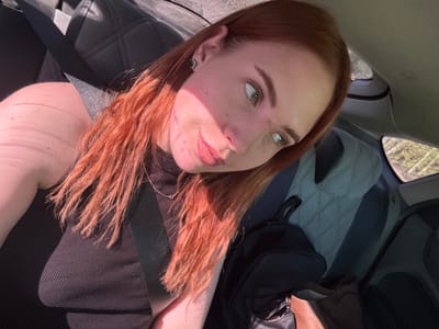 A woman with red hair and fair skin is seated in a car. She is wearing a black top and a seatbelt. A backpack is visible on the back seat. The car interior has black and gray quilted seats. A window is partially visible on the left side of the frame. The woman's face is partially illuminated by sunlight. Location: Likely within a car, no city context can be extracted. No connection to Starobaltachevo, Russia.