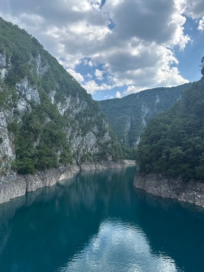 This is a vertical, high-angle, close-up shot of a body of dark teal water flowing between steep, rocky, and densely forested mountains. The mountainside on the left is covered in green trees and has a rocky texture. The water is calm, with ripples on the surface and a reflection of the sky, which is partly cloudy and blue. The image was taken on a bright, sunny day. The scene depicts a natural landscape, likely a river or a fjord, in Bezuje, Montenegro. There is no text visible in the image.