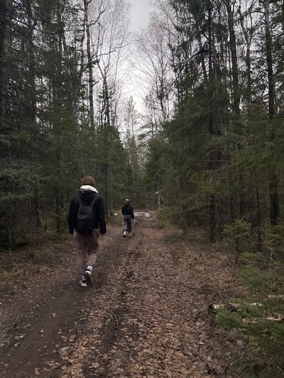 Two individuals walk along a dirt path covered in fallen leaves. The path is bordered by dense forest. The location is the city of Kachkanar, Russia. The first individual, closer to the viewer, wears a dark jacket, tan pants, and sneakers with white socks. They have a backpack on. The second individual, further down the path, is wearing darker clothing. The sky is overcast.