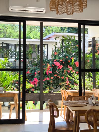 The image shows an indoor dining area with a view of lush greenery and buildings through a large glass sliding door. Inside, several wooden tables with woven placemats and chairs are visible. The tables are set with napkins and some appear to have price tags. In the foreground, a wooden chair with a woven back is prominently featured. A Hisense air conditioning unit is mounted on the white ceiling. Outside, a dense bush with vibrant pink flowers and green foliage is a focal point. The background reveals white buildings with dark-framed windows, surrounded by trees. Sunlight streams in, creating bright highlights on the tiled floor. The location is identified as Goron Talo, Indonesia.