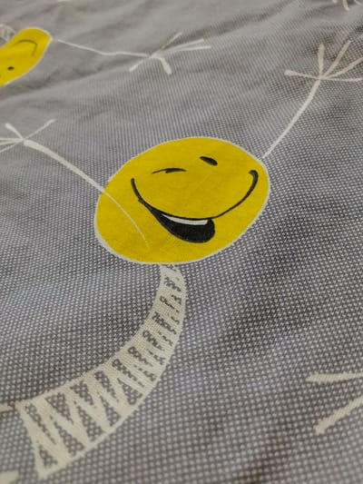 The image shows a close-up of a grey fabric with a pattern of yellow smiley faces and white dandelion-like shapes. One large, prominent smiley face is in the center right of the frame, with its mouth open and tongue sticking out. Another partial smiley face is visible in the upper left corner. The fabric has a textured appearance, likely due to its weave. There are no people or discernible elements of the environment or city of Zelenogorsk, Russia, in the image.