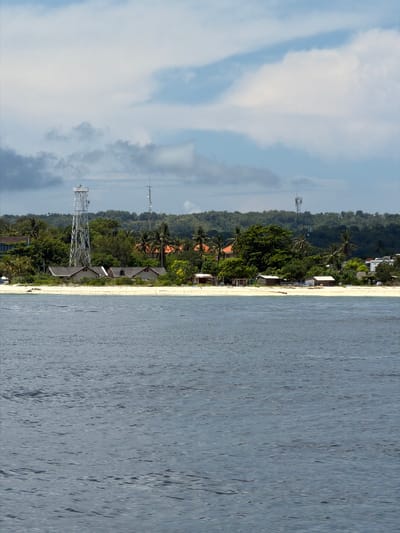 The image depicts a coastal scene in Jungutbatu, Indonesia. The view showcases a beach with light sand bordering a body of water, likely the ocean. The water is a pale blue-grey with ripples and variations in tone. On the land, a thin strip of beach is followed by a line of structures, appearing to be bungalows or houses, with dark-colored roofs. Behind these structures, dense vegetation dominates, including trees and shrubs. Interspersed within the vegetation are communication towers, tall metal structures. The sky is visible on the left side of the image, displaying a mix of pale blue and white clouds.