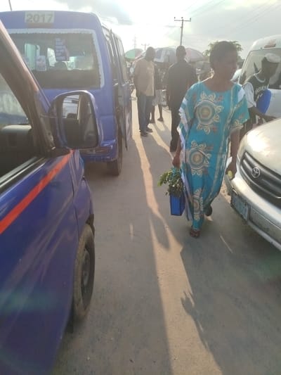 This media file captures a street scene in Uduagba, Nigeria, likely during a sunny day, as indicated by the strong shadows cast on the dusty road. The image features several vehicles, including a blue minivan with the number "2017" displayed on its windshield, and parts of other vehicles, one appearing to be a white van. The minivan has stickers on its rear window, one of which is partially legible and reads "GOD ALWAYS SHOWS UP ON TIME KEEP PRAYING," and another sticker with text including "DOES NOT EXIST."

In the foreground and mid-ground, several people are present. A woman in a light blue and white patterned dress is walking towards the right side of the frame, carrying a blue bag filled with green leafy plants in her left hand and a light-colored object in her right. Her expression is neutral as she walks alongside a white car. Other individuals are visible in the background, some standing and others walking, amidst what appears to be a bustling market or roadside area, with several umbrellas set up. The overall atmosphere suggests a typical day in a busy urban or semi-urban environment in Nigeria, possibly a market street or a transportation hub. The focus on the vehicles and the people engaged in daily activities provides a glimpse into local life.