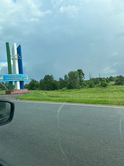 The image captures a roadside view in Irnykshe, Russia, featuring a prominent welcome sign. The sign is supported by three vertical columns, colored green, white, and blue, topped with a stylized, golden, starburst-like ornament. A blue banner across the lower part of the sign displays text in Russian, reading "АРХАНГЕЛ РАЙОНА" on the top line and "АРХАНГЕЛЬСКИМ РАЙОН" on the bottom line, indicating the Arkhangelsk region. The sign is anchored to a reddish-brown, stone-like base.
To the right of the sign, a verdant, grassy hill rises, densely populated with various trees. The sky above is overcast with heavy, grey clouds, suggesting an impending storm. In the foreground, a section of asphalt road is visible, with a white line marking the lane. The corner of a car's side mirror is visible in the lower left portion of the frame, reflecting a person's hand and arm. The overall impression is a rural landscape on a cloudy day, marking an entrance to the Arkhangelsk region.