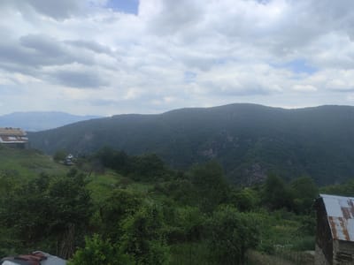 This media file depicts a scenic view of a mountainous landscape in Leshten, Bulgaria. The scene is dominated by lush green hills and mountains, covered in dense vegetation. The sky is overcast with dramatic, layered clouds, suggesting it might be late afternoon or early evening, or simply a cloudy day.
In the foreground, there are several objects and elements related to a rural setting. On the left, a rustic building with a weathered, corrugated metal roof is partially visible. Further down the slope, another building with a similar roof can be seen, nestled among the trees. A dirt path or road winds its way up the hillside, with a dark-colored vehicle parked on it. The vegetation is dense, with various trees and shrubs creating a textured, natural environment. A rough fence made of wooden posts and wire is also visible, delineating some of the areas. In the lower foreground, the edge of another roofed structure is visible, with some red and dark colored objects on it, possibly parts of vehicles or storage.
The overall impression is one of a remote, tranquil, and somewhat wild natural setting. The cloudy sky adds a sense of moodiness to the scene. There is no visible text in the image. The focus is on the natural beauty of the landscape and the rustic human presence within it.