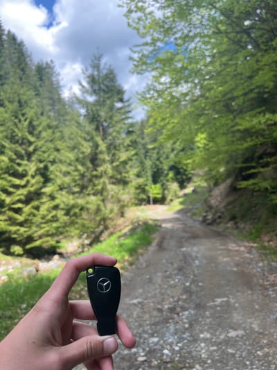 The image depicts a hand holding a black Mercedes-Benz car key in an outdoor setting. The background is a dirt road surrounded by green vegetation, likely a forest or a trail. The sky is visible through the trees, showing some clouds. The location is suspected to be near Drianovo, Bulgaria, based on contextual information. The primary objects are the car key, the hand, the dirt road, trees, and the sky. The action is someone holding the key, possibly contemplating a drive or having misplaced the car. There are no other people visible in the frame.