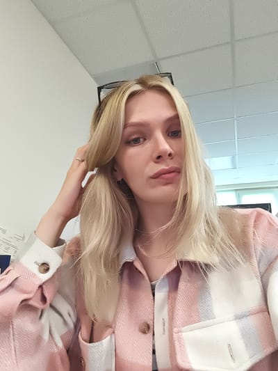 This is a medium shot, self-taken photograph of a young woman with blonde hair. She is looking directly at the camera with a neutral expression. Her hair is styled with some strands falling over her face, and she is touching her hair with her right hand, which has a ring on it. She is wearing a pink and white plaid shirt over what appears to be a striped t-shirt. There are glasses perched on top of her head. The background is an indoor setting with a white ceiling that has tiled panels and some fluorescent lighting. The walls are also white. To the left of the woman, there is a sheet of paper with visible text that is mostly illegible. The overall lighting suggests it is daytime.