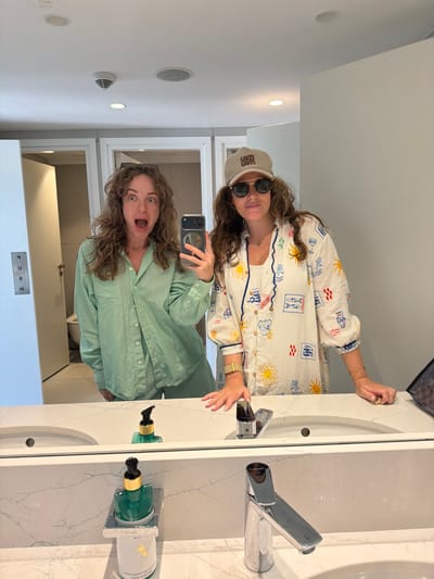 Two young women are taking a mirror selfie in a modern bathroom with white marble countertops. The woman on the left is wearing a mint green pajama set and has a surprised expression with her mouth open. She is holding a smartphone in her right hand, which is angled to capture the reflection. The woman on the right is wearing a white pajama top with a colorful print of suns, fish, and other nautical motifs. She has on sunglasses and a tan baseball cap. Her left arm is resting on the countertop, and her right hand is visible, showing a bracelet and rings. The bathroom features two sinks, chrome faucets, and soap dispensers. A "PUSH" sign is visible on a door in the background. The overall impression is that of a casual, personal moment shared between friends in a well-appointed private space.