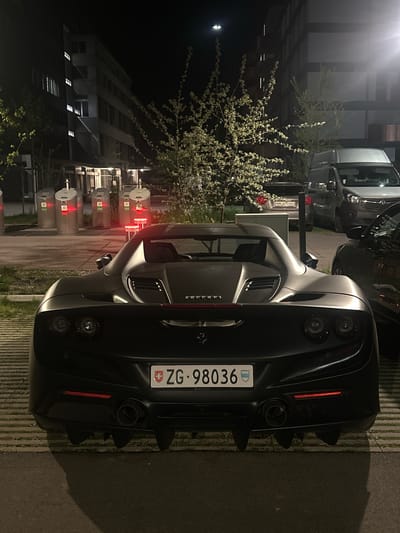 The image presents a dark, nighttime scene in Dübendorf, Switzerland, featuring a black luxury sports car, specifically a Ferrari, parked outdoors. The car is viewed from the rear, highlighting its sleek body, four circular taillights, and a prominent rear diffuser with quad exhaust pipes. A Swiss license plate, "ZG-98036," is centrally affixed to the car's rear, with the small text "THE LIVE CIRCLE" visible on its bottom edge. The "FERRARI" brand name is also visible on the trunk lid.
In the background, other parked vehicles are partially visible, including a light-colored SUV or van and a darker car. The scene is illuminated by artificial light sources, creating reflections on the cars and casting a glow on a nearby flowering tree or bush adorned with numerous white blossoms. Further in the background, dimly lit buildings with some illuminated windows are discernible against the dark night sky. The ground appears to be a paved surface, likely a parking area, with subtle striped markings. Several cylindrical, light-colored objects, possibly waste containers or urban furniture, are lined up to the left of the scene. The image captures a static, urban nighttime environment with the high-performance vehicle as its central subject.