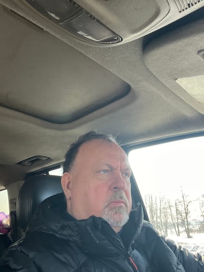 The image captures a middle-aged male seated inside a vehicle, facing and looking towards the right side of the frame. He has dark hair and a grey goatee, wearing a black puffer jacket with "NORTHLAND" visible on the left chest. The vehicle's interior features a light-colored headliner, an overhead console with a rectangular light fixture, and a visible sunroof opening. Through the vehicle's window, a bright, snowy landscape is visible, showing snow-covered ground and bare deciduous trees or shrubs under a bright sky, indicative of a winter season. The scene takes place in Ерино, Russia.