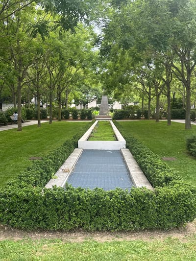 The image depicts a public park with manicured lawns, hedges, and a central monument. In the foreground, a rectangular pool area with a metal grate bottom is bordered by concrete and dense green hedges. Beyond this, a pristine green lawn stretches towards a monument at the far end of the park. The monument appears to be a tall, tapering obelisk or similar structure. A pathway lined with trees and hedges runs along the sides of the lawn. On the left side of the path, a person is seated on a bench, facing away from the camera. The park is surrounded by trees, some of which have full, green foliage, suggesting it is spring or summer. The architecture in the background is modern and appears to be part of the urban environment of Enghien-les-Bains, France. The overall scene conveys a sense of tranquility and order within a city setting.