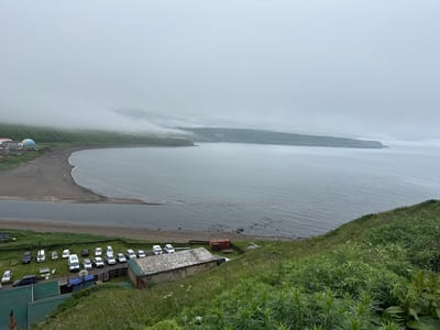 This image shows a coastal landscape in Kuril'sk, Russia, under a heavy overcast sky. In the foreground, a steep, grassy slope leads down towards a shoreline. Below the slope, a collection of buildings with green corrugated roofs and some weathered industrial structures are visible. A dirt road runs along the edge of the buildings, and a parking lot with several cars is situated next to it.

The shoreline itself is a mixture of sand and pebbles, bordering a calm, grey body of water. Further out, the water stretches towards a distant, rolling coastline covered in green vegetation and shrouded in low-lying fog. A curved sandbar or spit of land extends into the bay. On the far left of the image, a cluster of buildings includes a distinctive geodesic dome structure. The overall impression is one of a remote, somewhat industrial coastal settlement on a cloudy day.