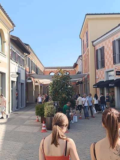 The image depicts an outdoor pedestrian area in Serravalle Scrivia, Italy, possibly a shopping outlet or commercial district. The street is paved with light gray bricks and flanked by multi-story buildings with pastel-colored facades. Some buildings feature dark brown shutters, while others have awnings over storefronts. Store signs, including "TWINSET" and "BALLY," are visible.
A small gathering of people are scattered along the street, some walking, others standing or sitting. Traffic cones are placed near planted shrubs and a tree in the center of the walkway. In the foreground, two women are facing away from the viewer, with a partial view of their backs and ponytails. The woman on the right has a tattoo on her back. The weather appears sunny with a clear, blue sky.