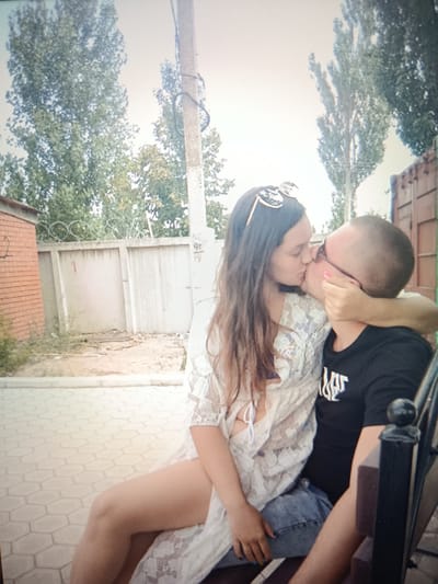 In an outdoor setting, a young couple is captured in a passionate embrace, their lips locked in a kiss. The woman, with long, flowing brown hair, wears a white, sheer lace cover-up over a bikini top, and her sunglasses are perched atop her head. The man, with a shaved head and glasses, is dressed in a black t-shirt. They are seated side-by-side on a bench, the woman's arm wrapped around the man's neck. The background features a brick building on the left, a concrete fence with barbed wire, and trees. The ground is paved with hexagonal tiles. The image appears to be taken in the city of Pishchane, Ukraine, suggested by the visible Cyrillic writing on a utility pole and a red shipping container in the background, which is characteristic of industrial areas found in such locations.
