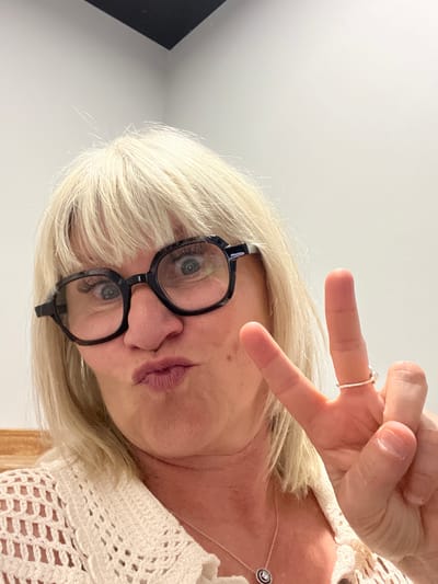 An angled, close-up indoor photograph depicts an adult, likely female, in Saint-Priest-en-Jarez, France. The subject has short, bright blonde hair and wears black-framed glasses, through which her blue-grey eyes are visible, reflecting a light source. Her lips are pursed in a slight pout. She is wearing a light-colored, possibly cream or off-white, crocheted top, and a thin silver necklace with a small, circular pendant adorns her neck. With her right hand, she forms a peace or victory sign, with a silver ring on her index finger. The background is a plain, bright white or off-white wall or ceiling, indicating an uncluttered indoor setting with clear, even lighting. The overall impression is one of playfulness or lightheartedness. No text is visible in the image.