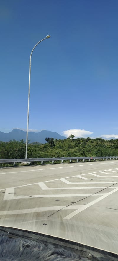 The image depicts a clear blue sky over a landscape in Lambarotunong, Indonesia. A tall, silver streetlight stands on the right side of a paved road. The road has white lane markings and appears to be an elevated highway due to the guardrail. Beyond the guardrail, a dense green forest covers the land, with rolling hills and mountains in the distance. A single white cloud is visible in the sky above the trees. The foreground shows a portion of the vehicle from which the image was taken, including a fluffy, dark blue seat cover.